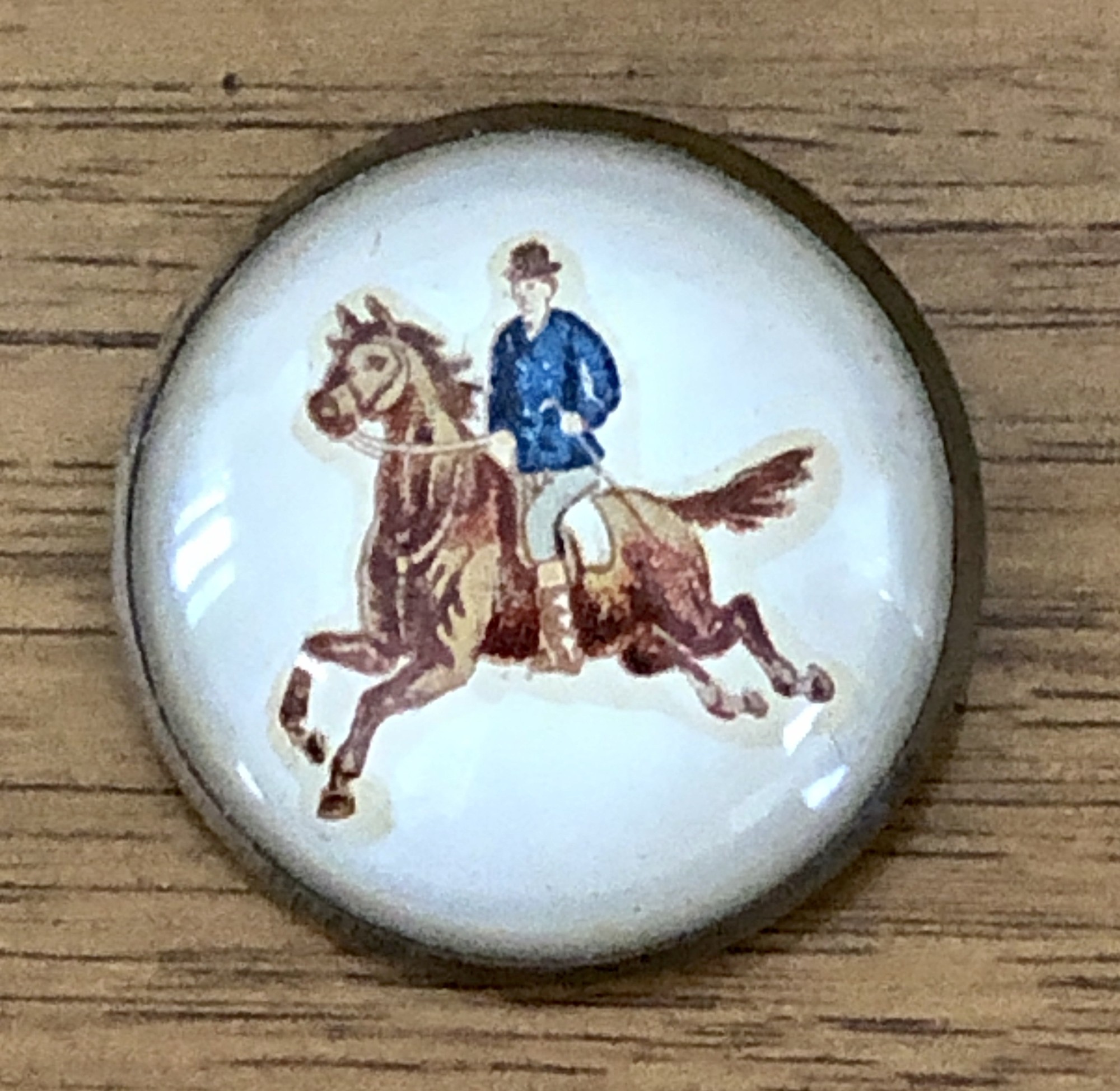 Victorian c.1980-1910 Domed convex glass horse Bridle Rosette Brooch.
Galloping out of a creamy off-white background, this spirited galloping horse and rider are a Victorian die-cut made as a bridle rosette brooch style pin. At 1-5/8 inches across, the clear artwork is an eyecatcher. The back of the rosette was designed to slide a horse bridle strap through.