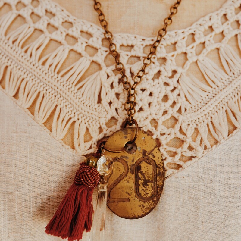 This hand crafted necklace hangs on a 22 inch chain. It features an oval, brass tag with a 20 on it as well as a crystal and tassle.