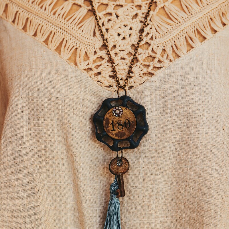 This unique necklace's pendant is a blue faucet knob with a brass plate engraved with 180. It also includes a brass key and blue tassle. It is on a 16 inch chain.