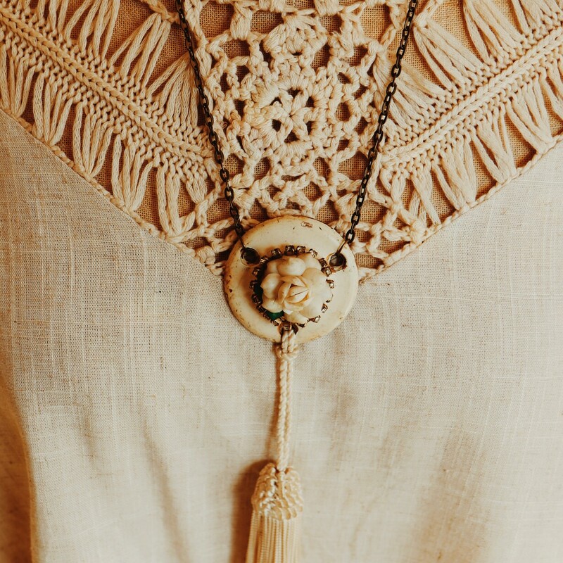 This hand crafted, one of a kind necklace has an antique door piece as its pendant with a flower on the front. From this hangs a white tassle. This piece is on a 23 inch chain.