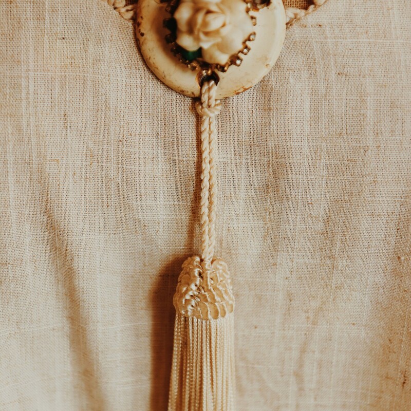 This hand crafted, one of a kind necklace has an antique door piece as its pendant with a flower on the front. From this hangs a white tassle. This piece is on a 23 inch chain.