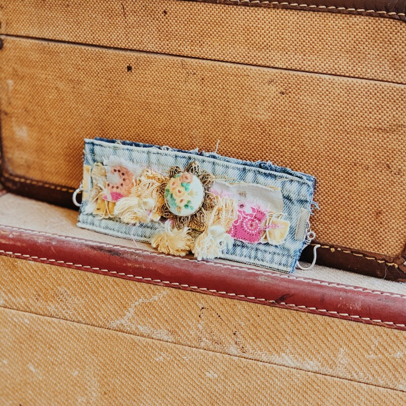 This fabulous, hand crafted denim cuff by Kelli Hawk Designs measures 8 inches in length and has a hook and eye clasp. The artist used a collection of vintage fabrics and charms to create a one of a kind centerpiece!