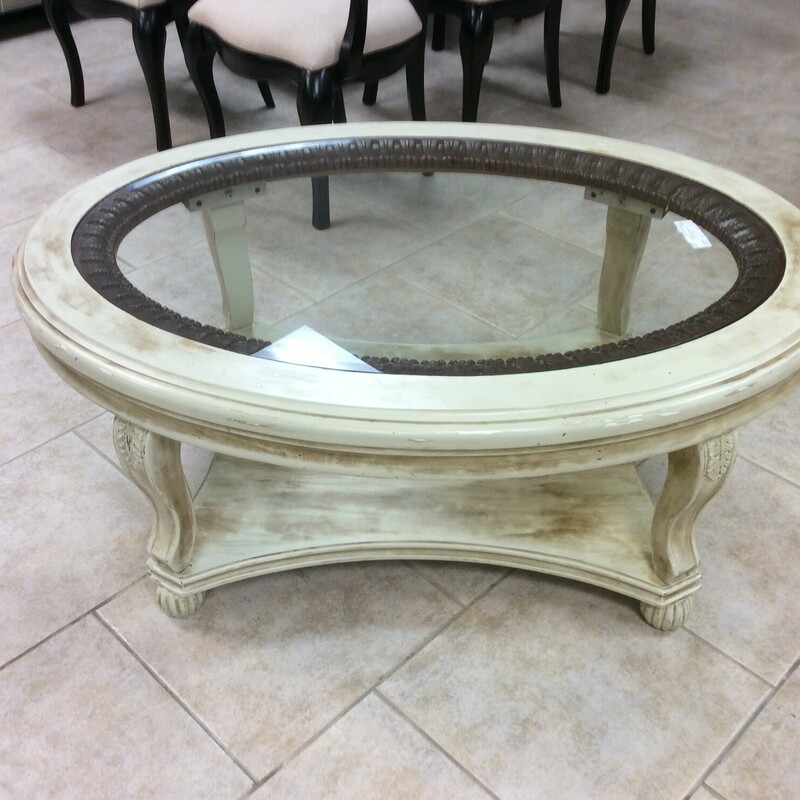 This is a antique distressed cream oval coffee table. This coffee table has a oval glass top and brown leaf detail.