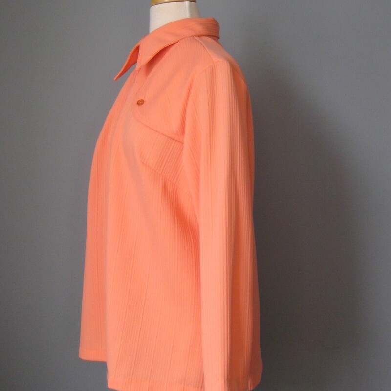 Fun 1970s buttondown in bright orange.  (it looks a little peach in my photos but it is more orange than peach)<br />
Made of ribbed poly double knit (tiny bit of stretch)  it's very simple with a little flap/button detail at the shoulders which gives it a hint of western flair.<br />
plain sleeves<br />
no tags (they were removed at some point)<br />
unlined<br />
great shape.<br />
<br />
flat measurements:<br />
shoulder to shoulder: 14.75 ( this is a little narrower that average)<br />
armpit to armpit: 21.5 (roomy here - gives the shirt a little boxiness)<br />
waist area: 23.5<br />
length: 26.25<br />
underarm sleeve seam: 15.5<br />
<br />
thanks for looking!<br />
#44985
