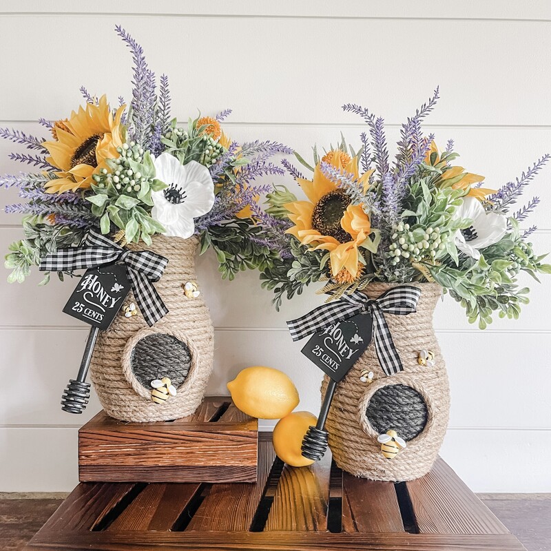 Our honeypot vase is the perfect centerpiece or table decor for your summer home decor! This adorable piece is hand-wrapped using jute rope around a glass vase to create the beehive look; and includes soooo many extras!!

The florals are all top quality faux pieces that are hand selected for the arrangement and include flocked lavender; golden sunflowers; yellow thistle and more! We love details; so we added the handmade honey sign and distressed honey dipper as accents along with the adorable bees and dainty buffalo check bow.

This piece measures approximately 16' tall from the bottom of the vase to the tip of the tallest floral stems; and approximately 8' wide/deep (florals - base is about 4').