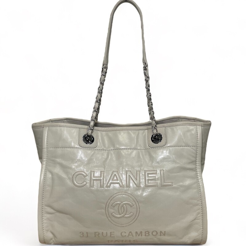 CHANEL Glazed Calfskin Small Deauville Tote in Ecru. This tote is crafted of fine glazed calfskin leather, with a Chanel advertisement logo stitched on the front, a diamond-quilted rear pocket, and caviar leather trim. The bag features leather-threaded polished silver chain link shoulder straps, and a wide top, open to a grey fabric interior with zipper and patch pockets.

Date code: 22311999
Year: 2016
Dimnesions:
Base length: 13.50 in
Height: 10.00 in
Width: 6.75 in
Drop: 10.00 in