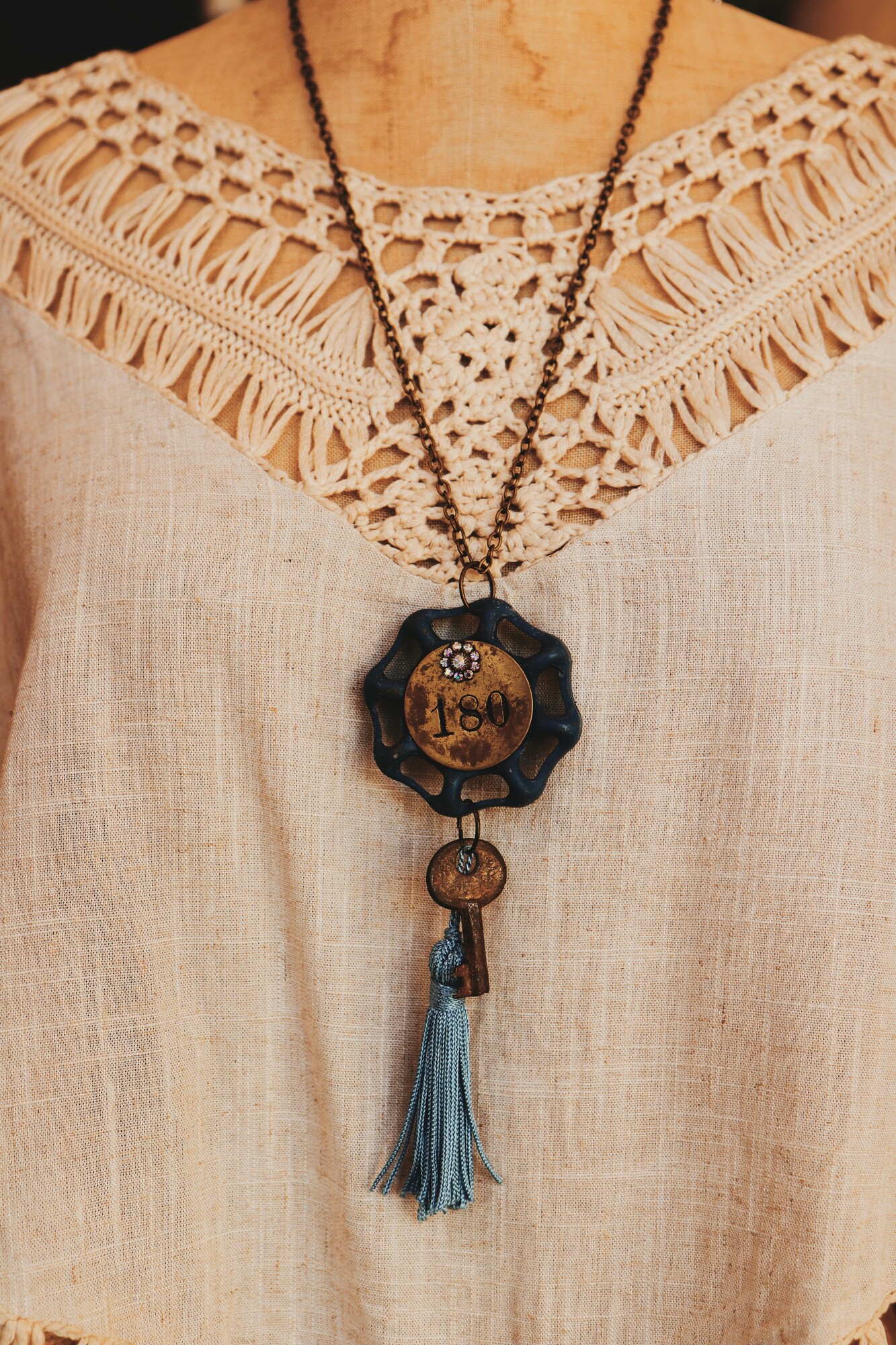 This unique necklace's pendant is a blue faucet knob with a brass plate engraved with 180. It also includes a brass key and blue tassle. It is on a 16 inch chain.