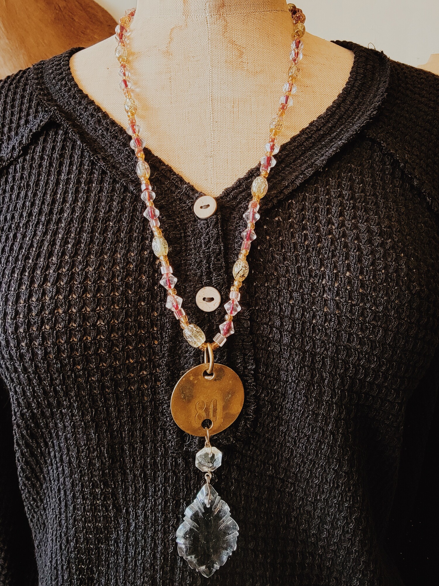 This one of a kind necklace is on a strand with a variety of pink, clear, and yellow beads. It has a circle brass plate as a pendant with 80 engraved in it and a crystal hanging from that. The necklace strand is 28 inches.
