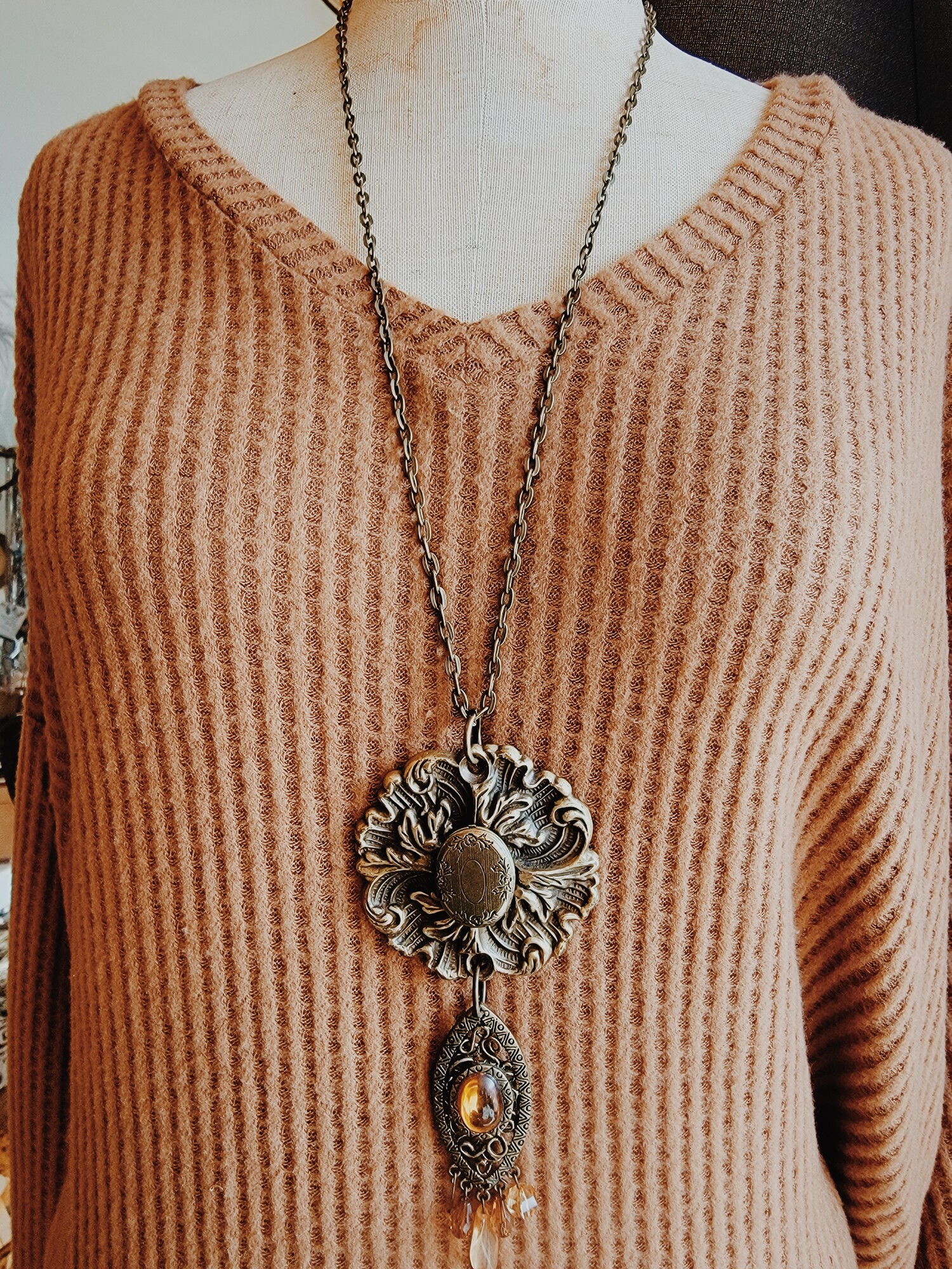 - Hand crafted necklace
- 30 inch chain
- Brass flower pendant with locket center
- Brass charm hanging from pendant with orange tone stones