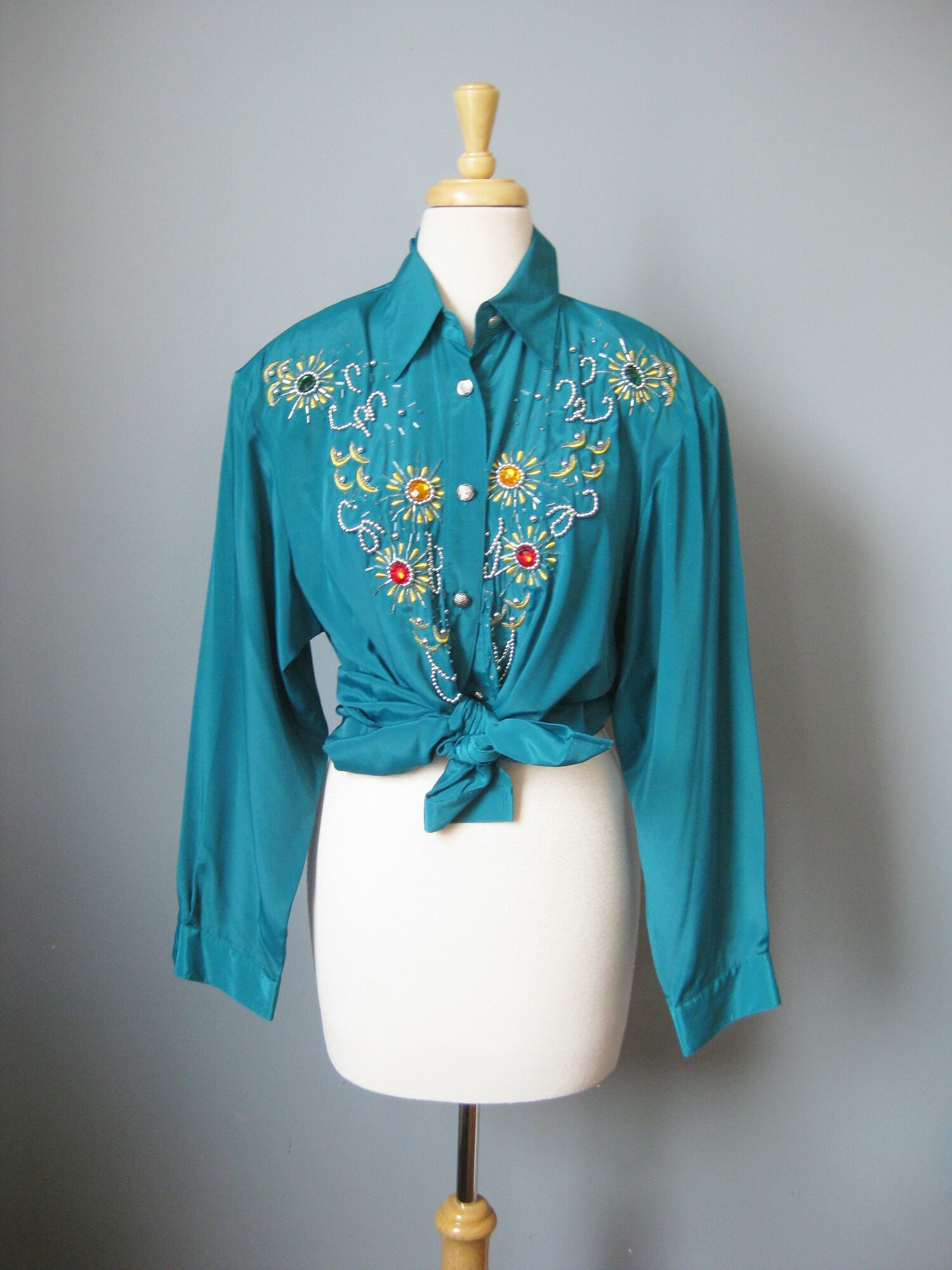 This long sleeved teal green shirt is lavishly bejeweled with large jewel tone stones  and gold beaded embroidery.

This piece was made in Hong Kong by Harbour Vue.   It's made of a slick matte polyester. The jewels are multi color plastic.  Good quality fabric in excellent condition.  Shoulder pads.

It is marked size 16, which I believe is pretty true to size today, but definitely use the measurements provided as your ultimate guide to fit.

These  are the flat measurements, please double where appropriate:
Shoulder to shoulder : 18
Armpit to Armpit: 21
Width at hem: 23
Underarm sleeve seam: 22
Length: 29 1/2

Thank you for looking.
#43094
